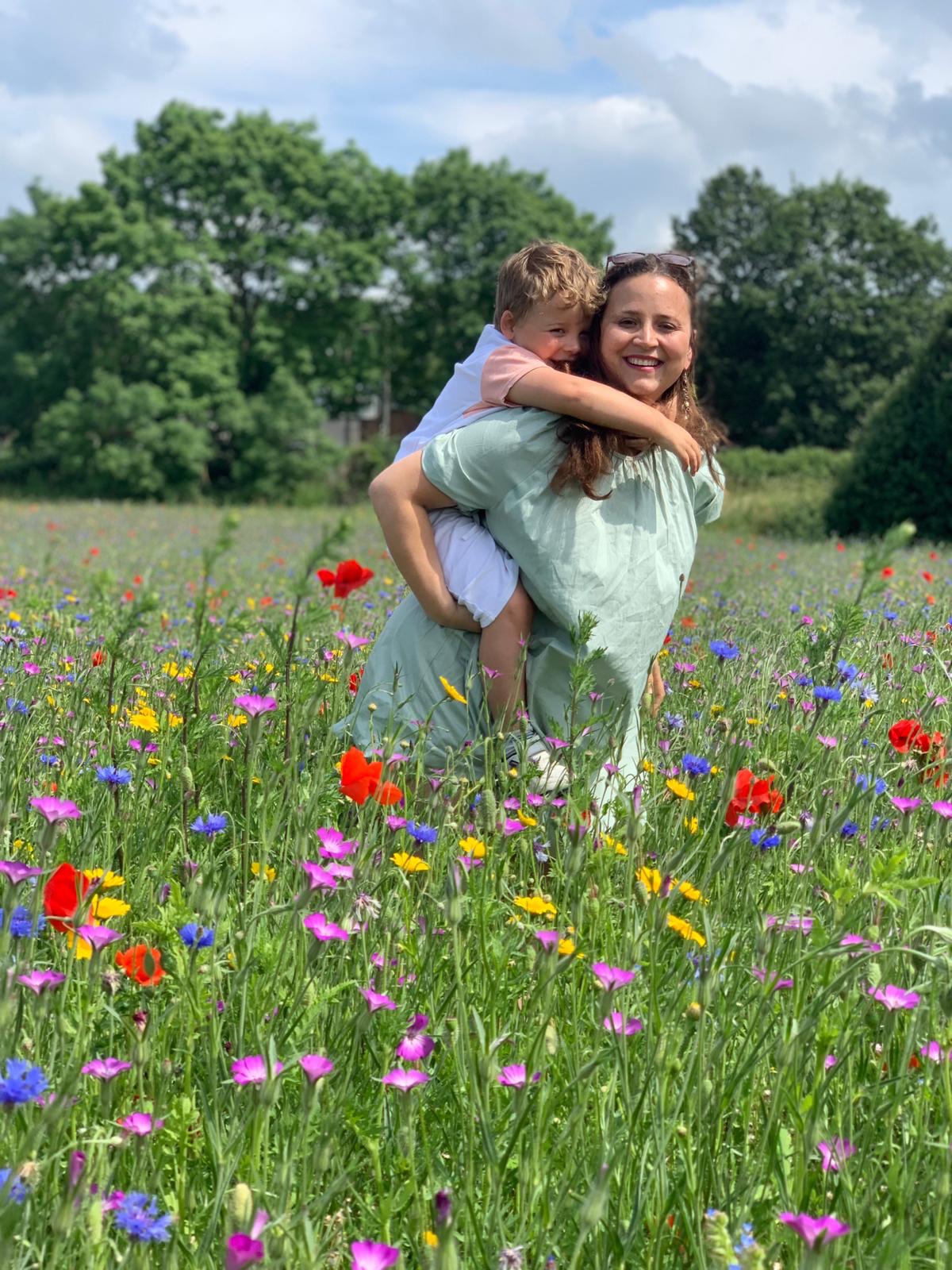 Lavania carrying Arlo on her back in the midst of a wildflower field on a sunny day.