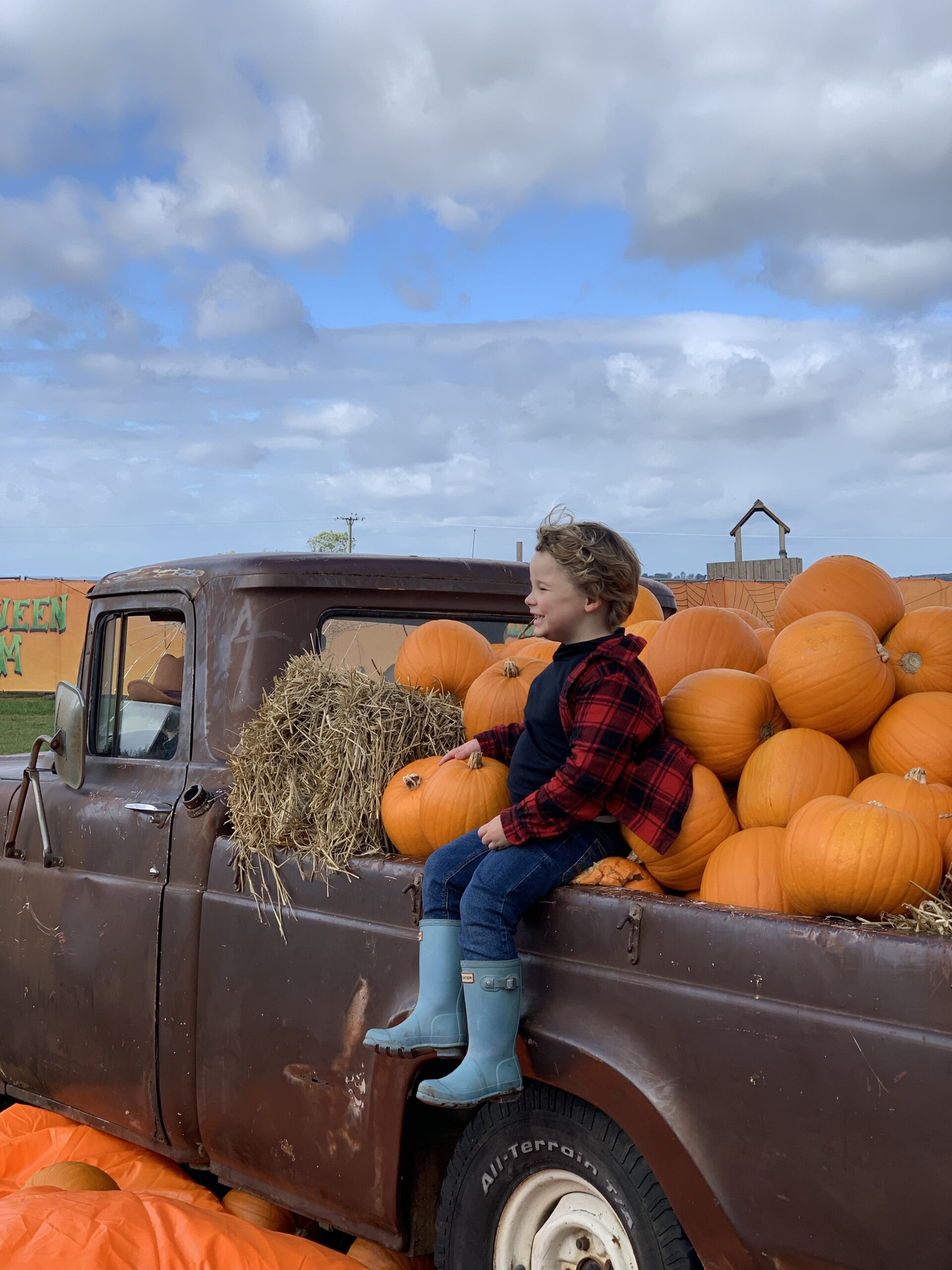 Blonde boy in a plaid shirt and jeans sits in a red truck filled with bright orange pumpkins and hay. He is wearing blue denim jeans and his legs dangle over the side - he is also wearing sky blue wellies. He is smiling with a grin and his curly hair is blowing in the breeze. Taken at Essington Farm - a childrens farm near the west midlands 