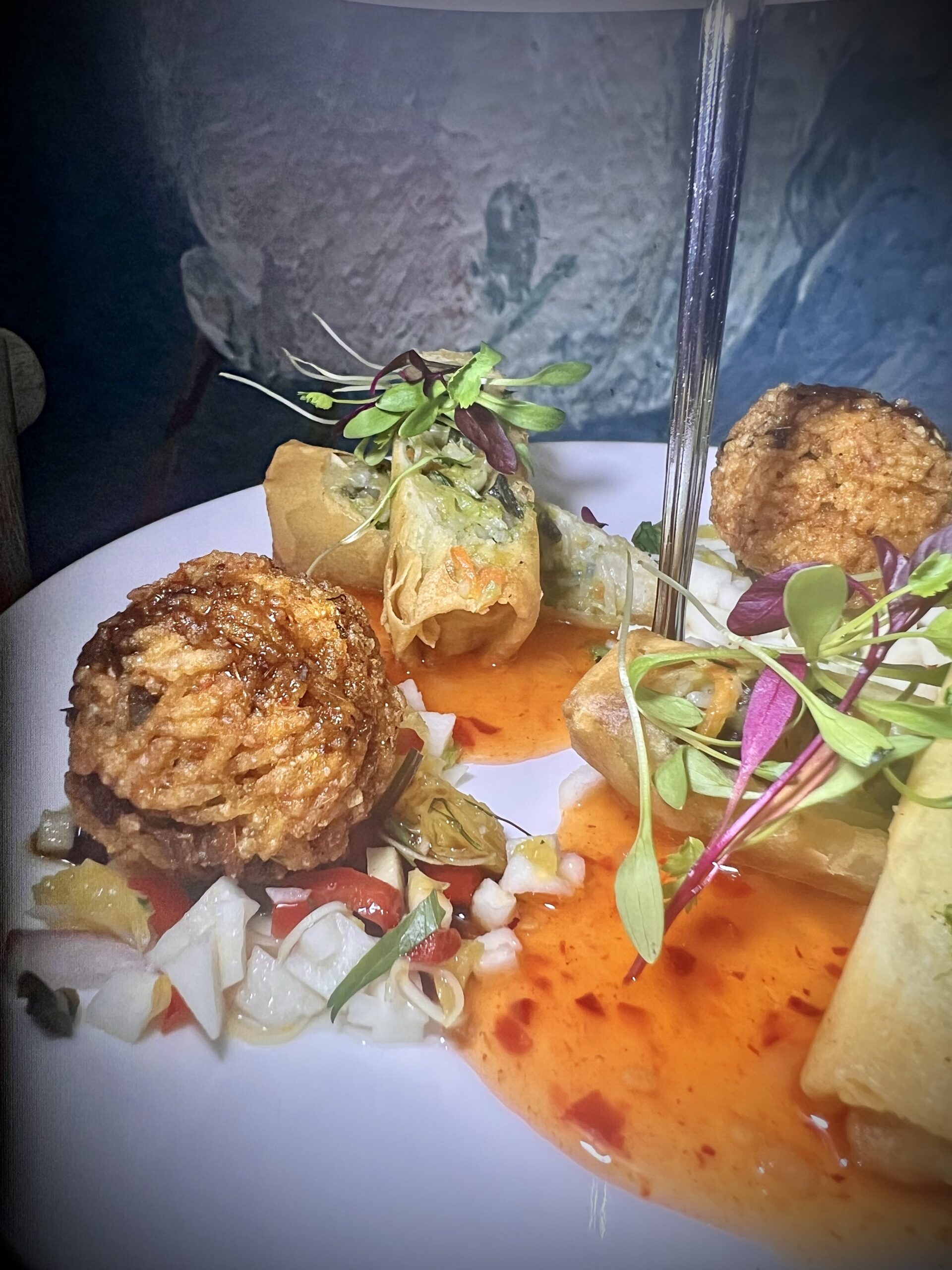 Plated gourmet meal from Giggling Squid with risotto balls, spring roll, and garnished with microgreens, served with a drizzle of red sauce.