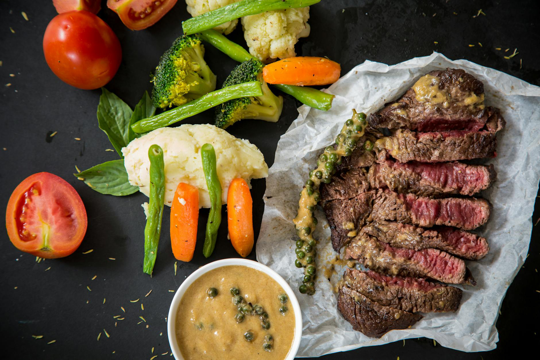 Sliced steak with peppercorn sauce accompanied by an assortment of steamed vegetables on a dark background.