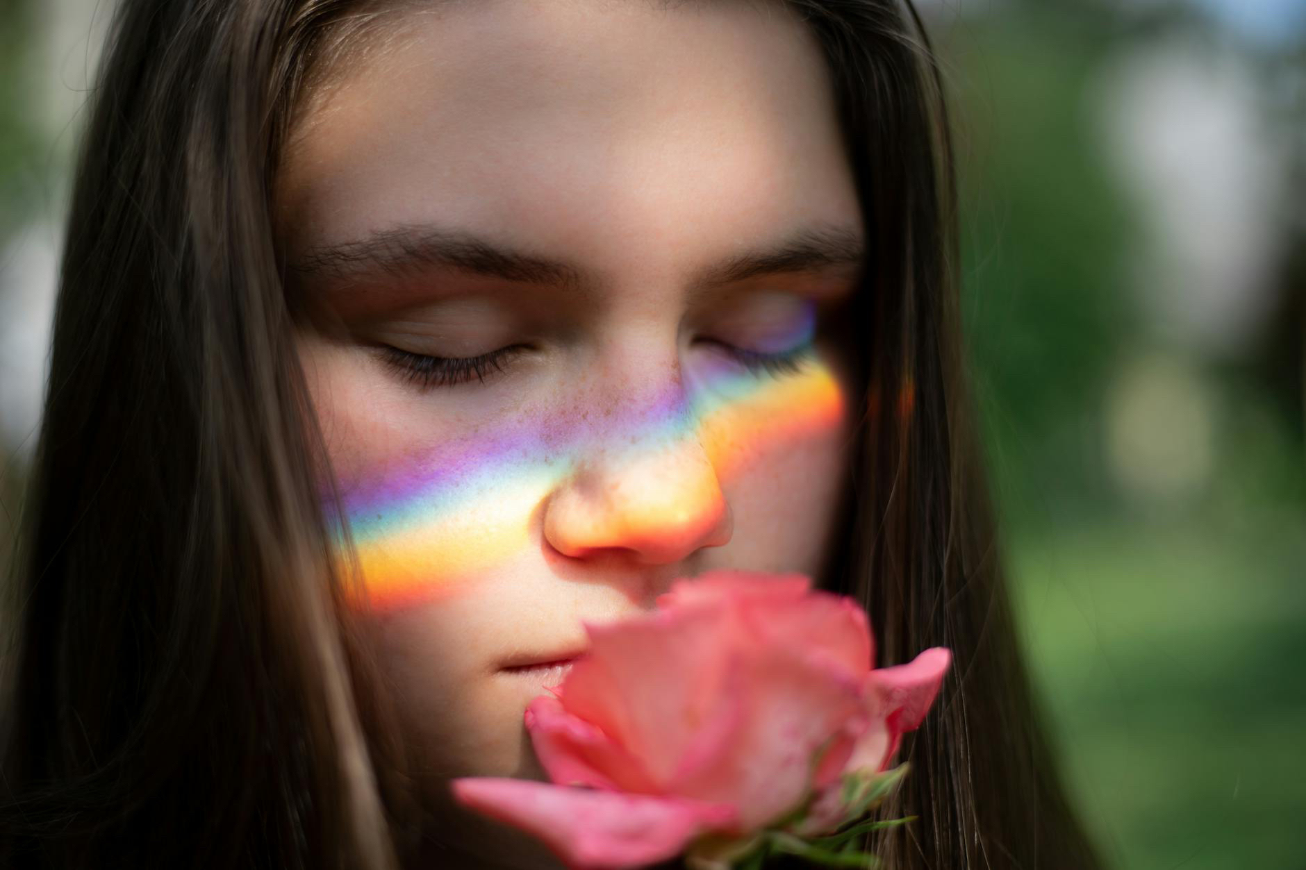 A girl with closed eyes smelling a pink rose, with a rainbow light spectrum reflected on her face.