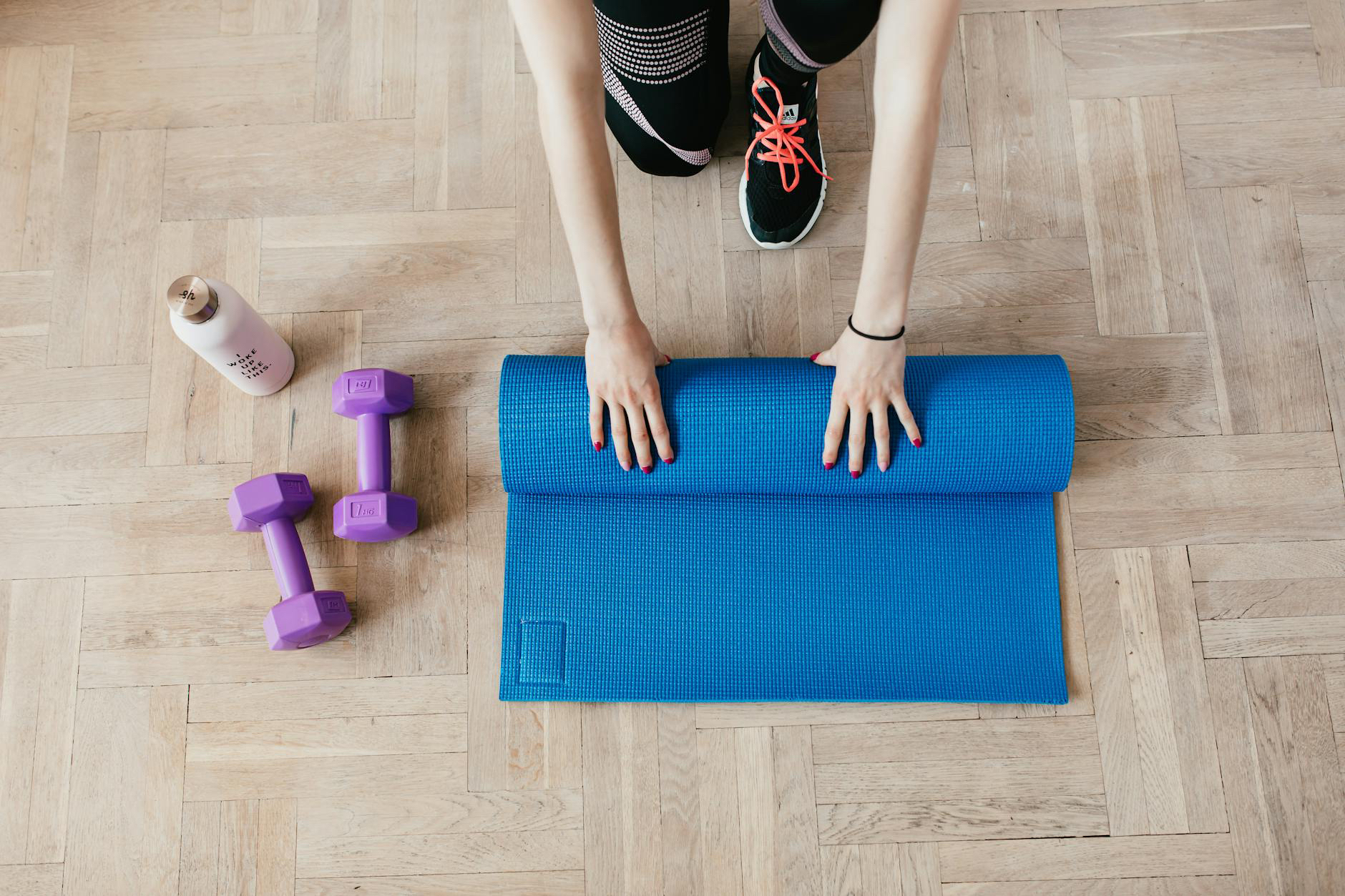 Woman rolling up a blue yoga mat on a wooden floor with purple dumbbells and a water bottle beside her.