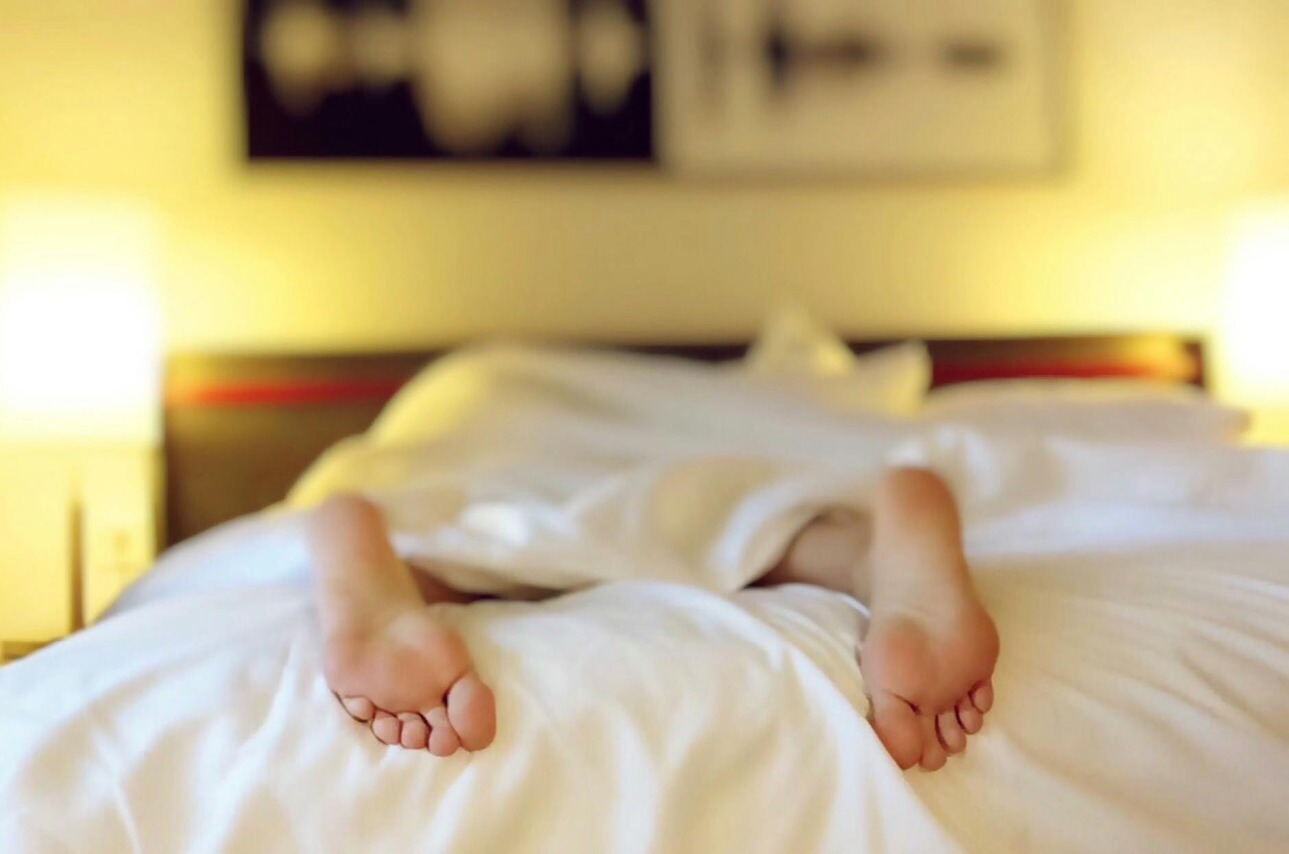 A person lying in bed with their feet sticking out from under a white blanket, in a softly lit room with two bedside lamps and abstract art on the wall.