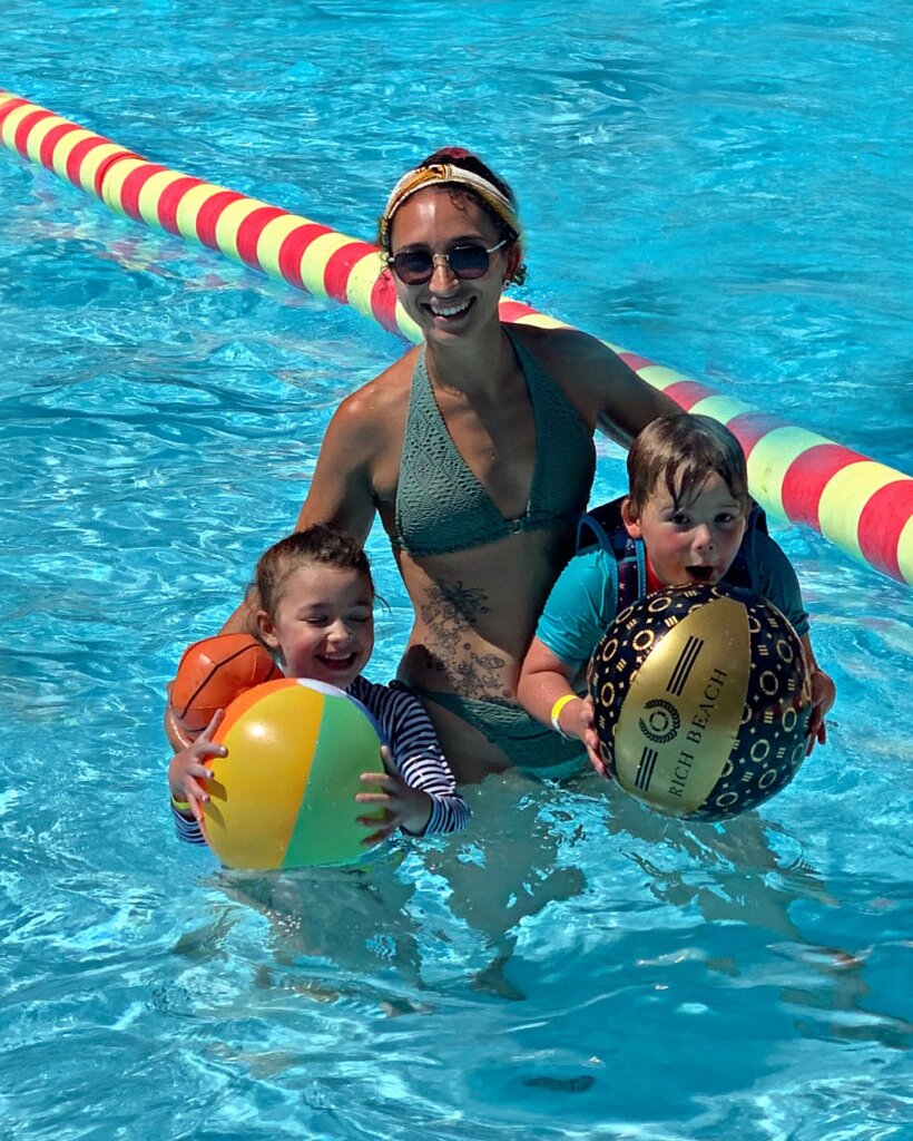 A woman in a green swimsuit stands in a pool, smiling, while holding two young boys and colorful beach balls. Red and yellow lane dividers are visible in the background.