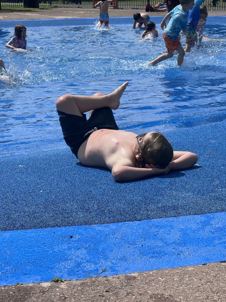 Child in black swim trunks and goggles lounges on a blue playground surface while children play in shallow water in the background.