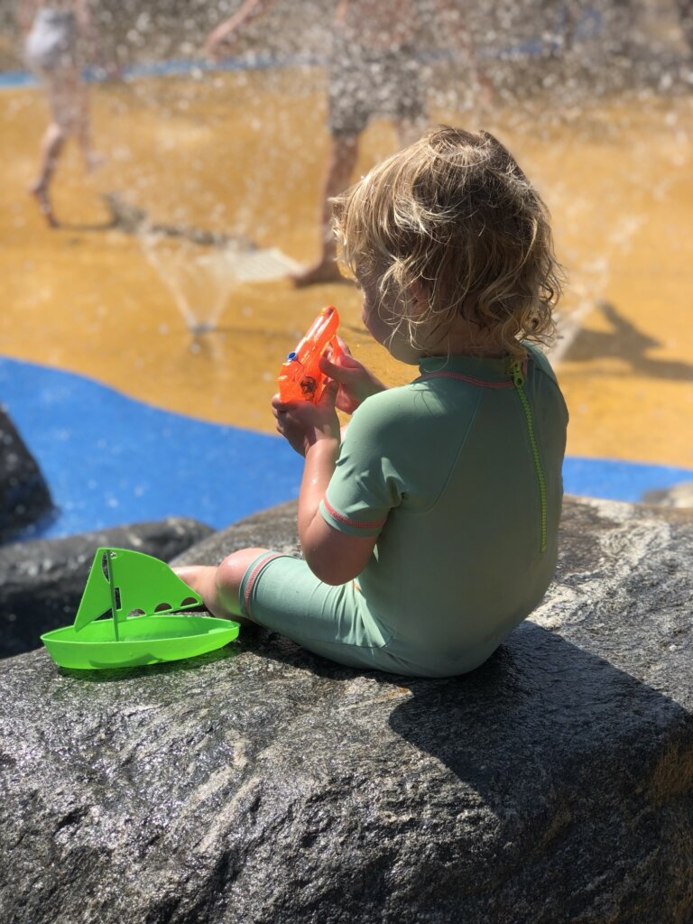 A child in a green swimsuit sits on a rock, holding an orange toy. A green toy boat sits beside the child. Water splashes in the background.