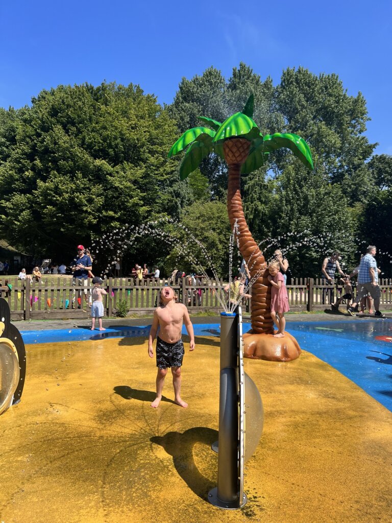 A child stands under sprayers at a water play area, with a large artificial palm tree and other children playing in the background on a sunny day.