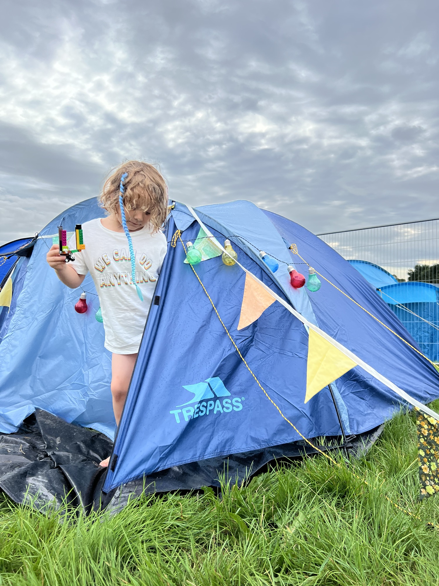 A child with a short hairstyle stands outside a blue tent decorated with string lights and bunting, holding colorful ribbon sticks. The sky is overcast, and the area has several other tents.