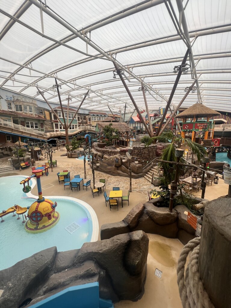 Indoor water park featuring a shallow wading pool, various rides, play structures, and seating areas under a translucent roof.