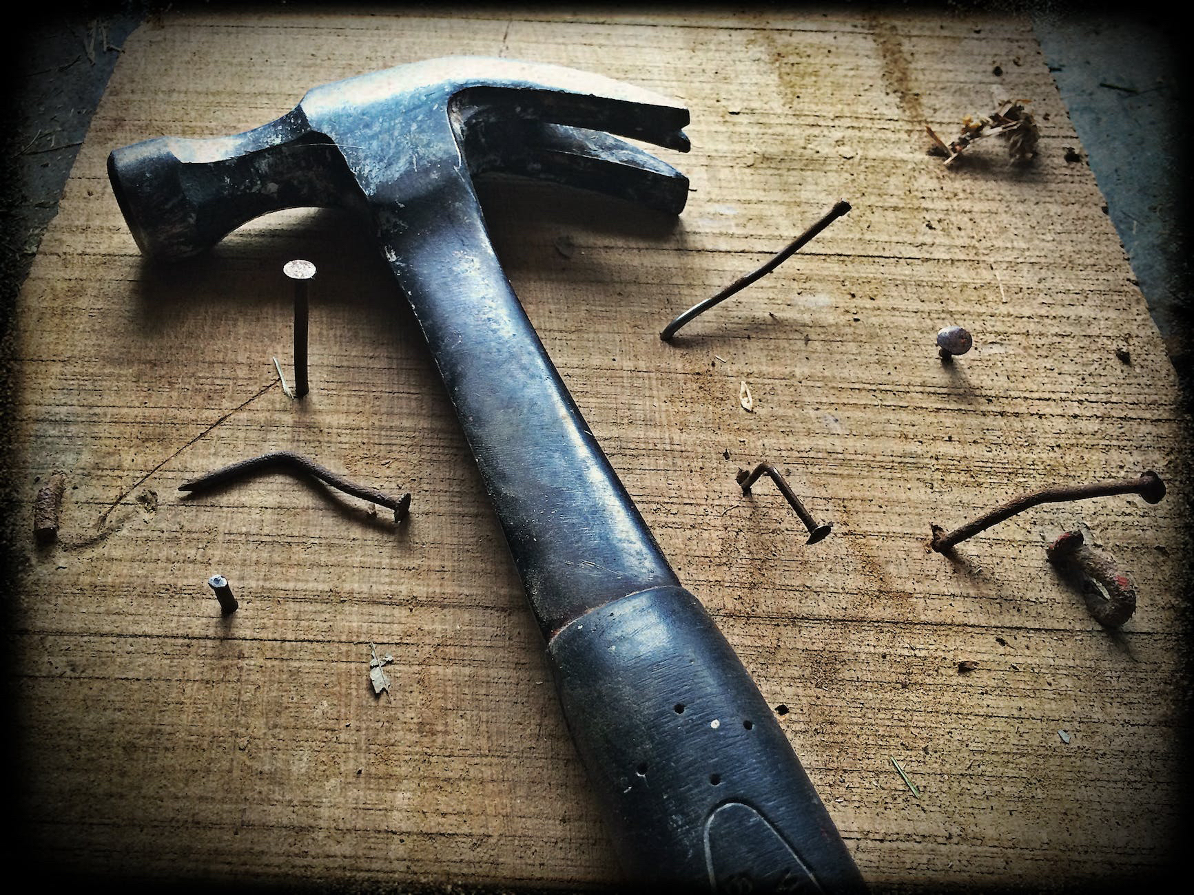 A hammer rests on a wooden plank surrounded by several bent and discarded nails.