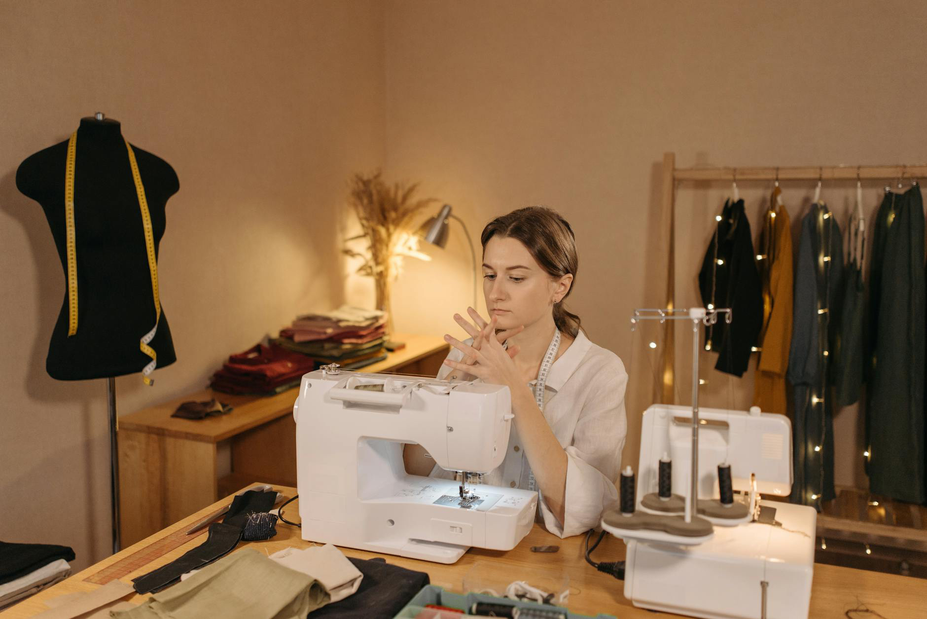 A person is seated at a sewing machine, with a mannequin and hanging clothes in the background, focusing on their hands.
