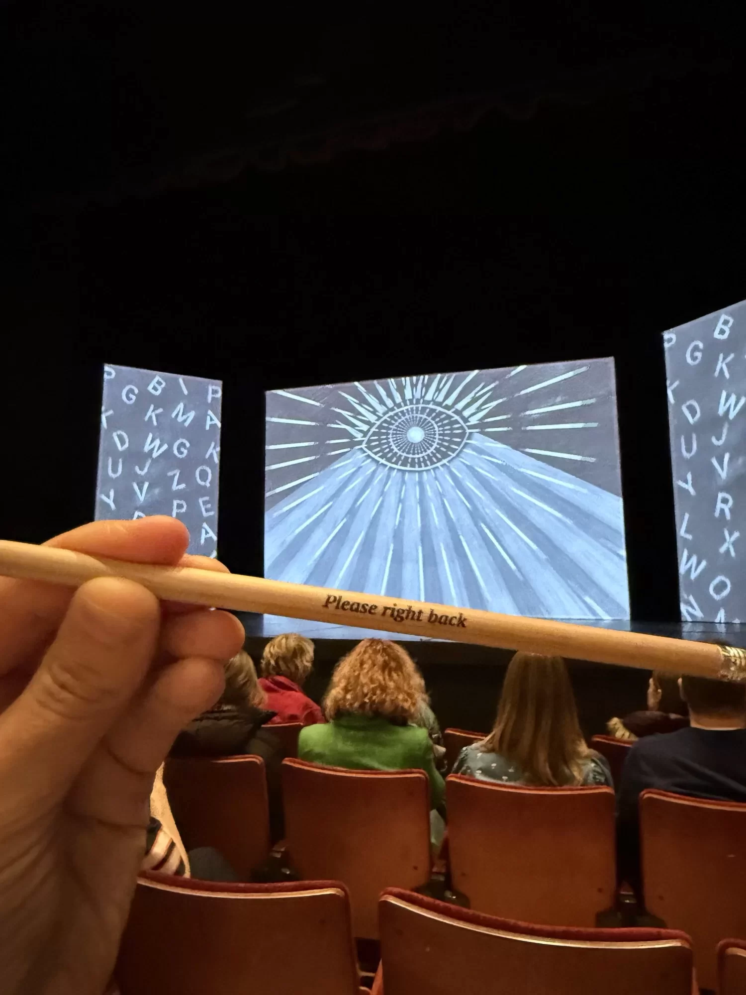 Hand holding a pencil with "Please fight back" text. Audience seated in a theater; stage displays abstract eye art and letters.