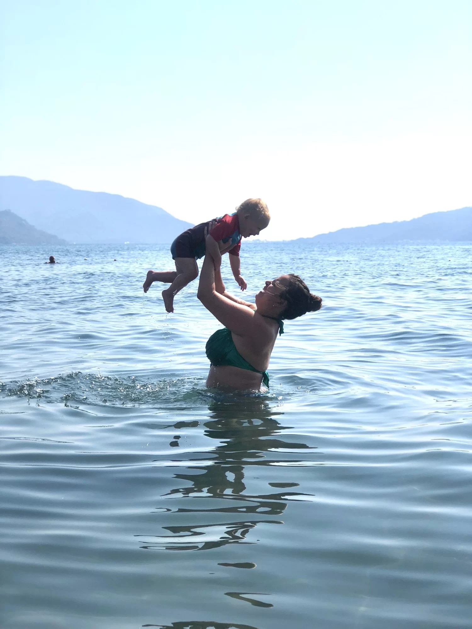 An adult lifts a child in the air while standing in a large body of water, with mountains visible in the background.