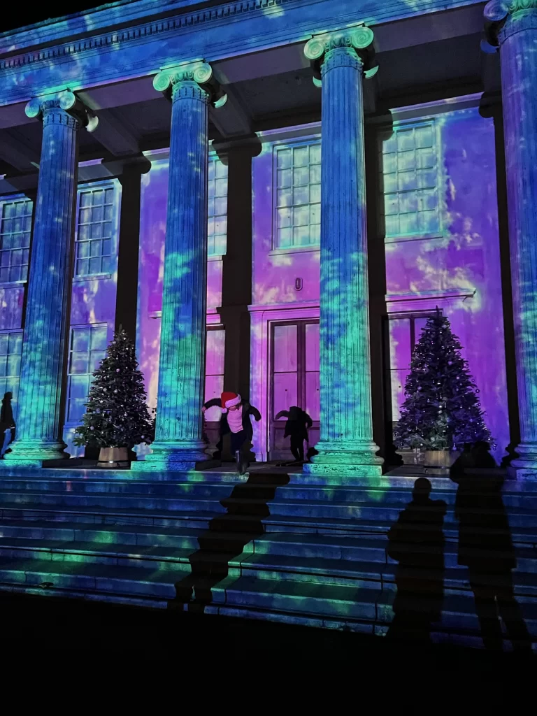 Building facade with illuminated projections of clouds and sky. Four large columns and several Christmas trees line the stairs, with silhouettes of people casting shadows.