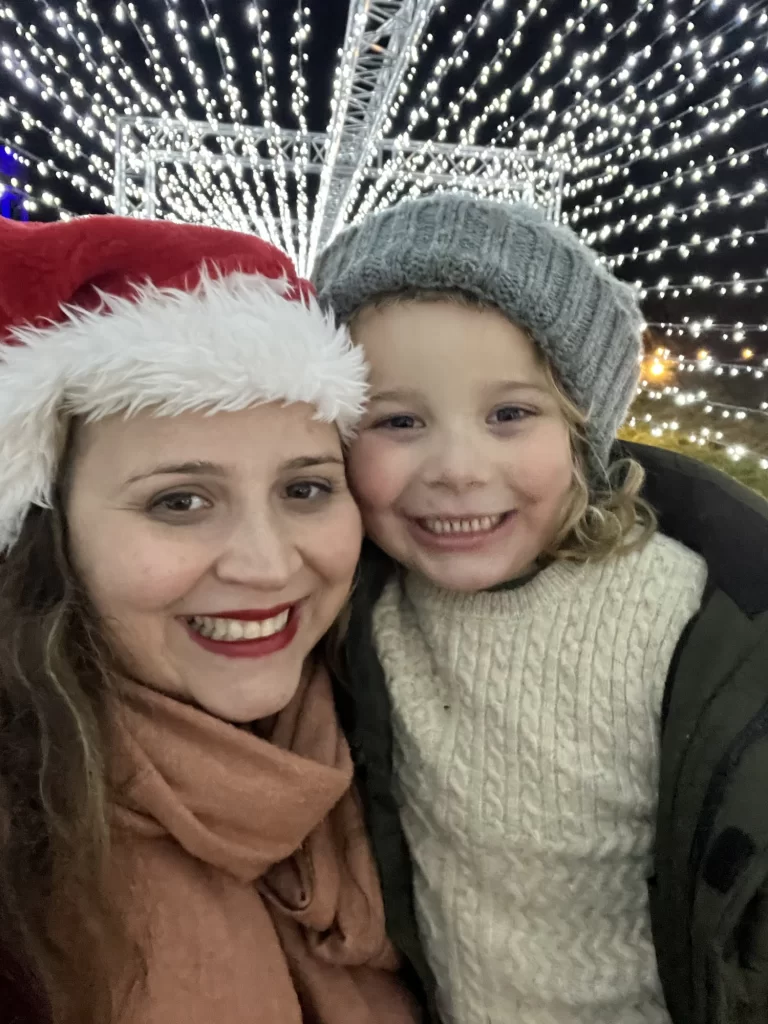 A woman in a Santa hat and a child in a gray beanie smile in front of a backdrop of twinkling lights.