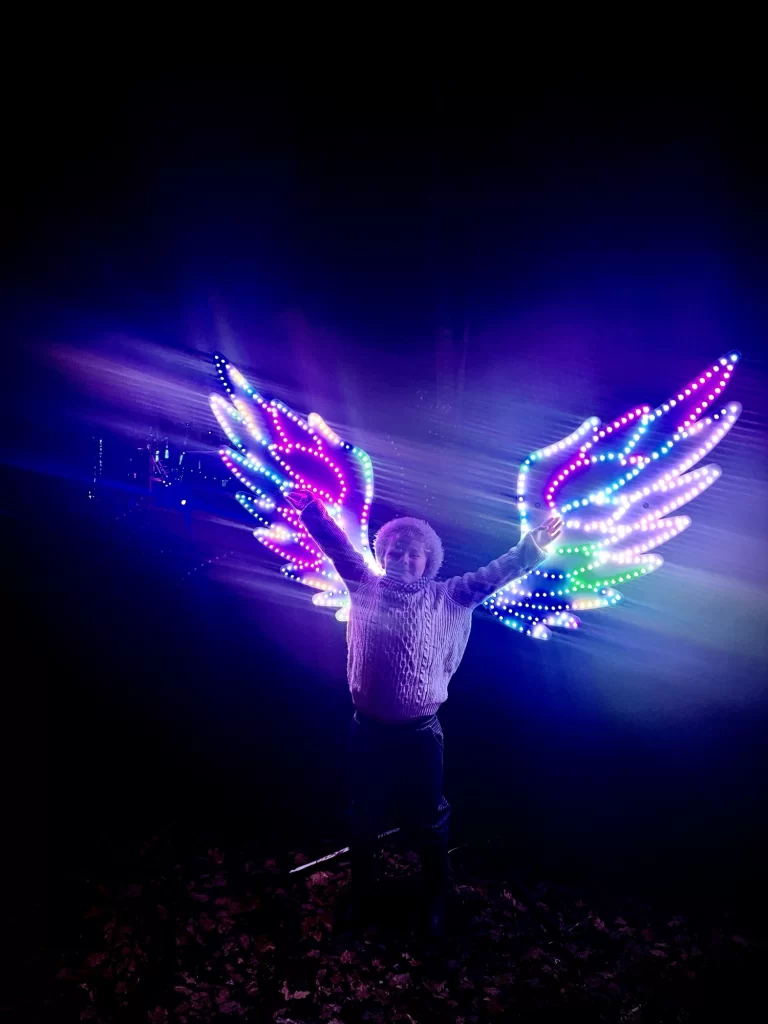 A person standing at night wearing glowing LED wings with colorful lights.