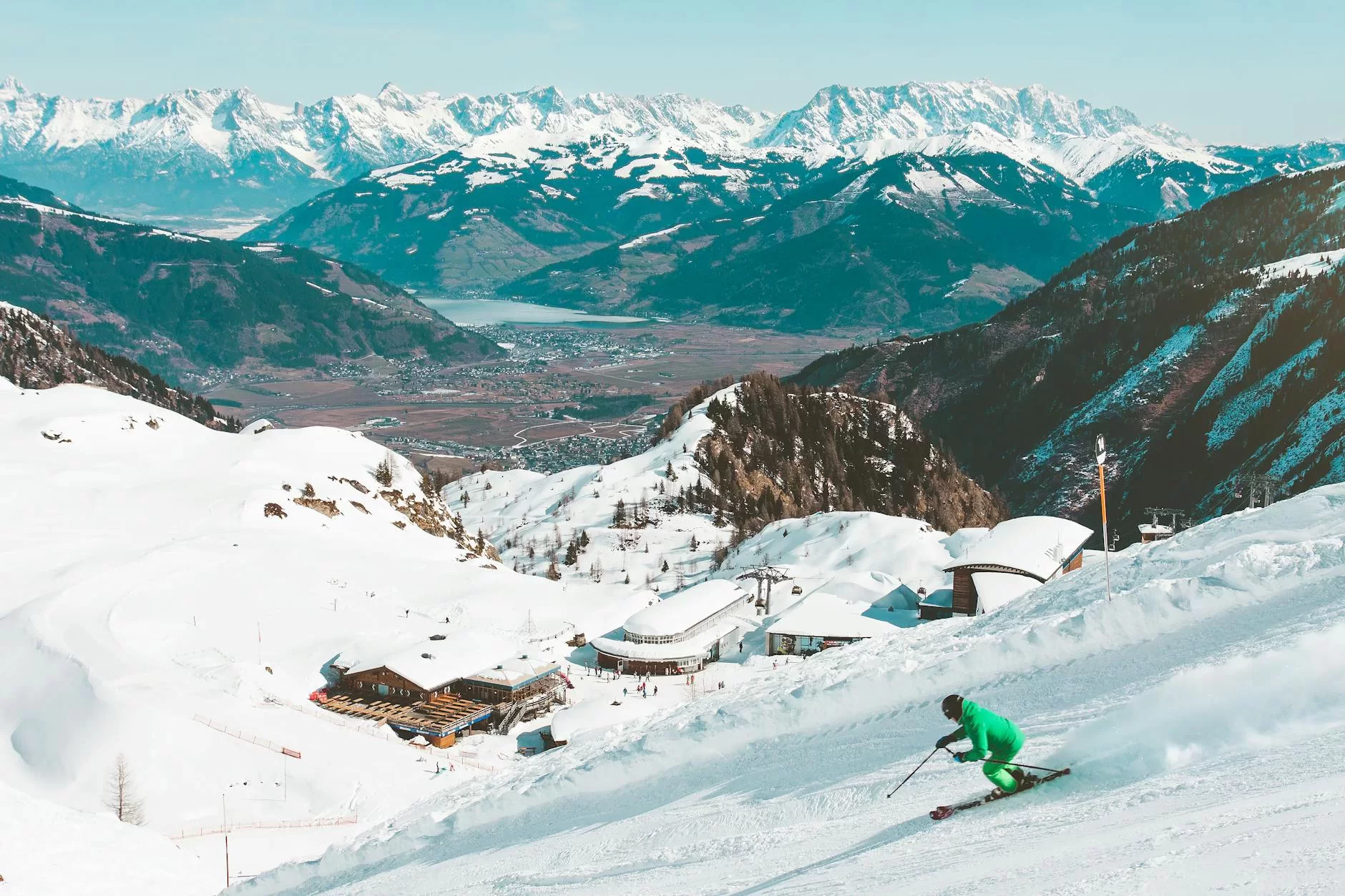 A skier in green descends a snowy slope with mountain peaks in the background on a clear day.