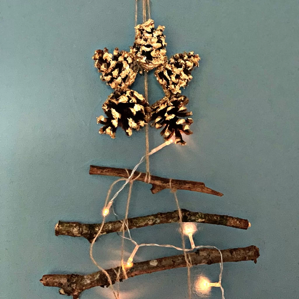 Pinecone star topper and tree made of sticks adorned with string lights, against a blue background.