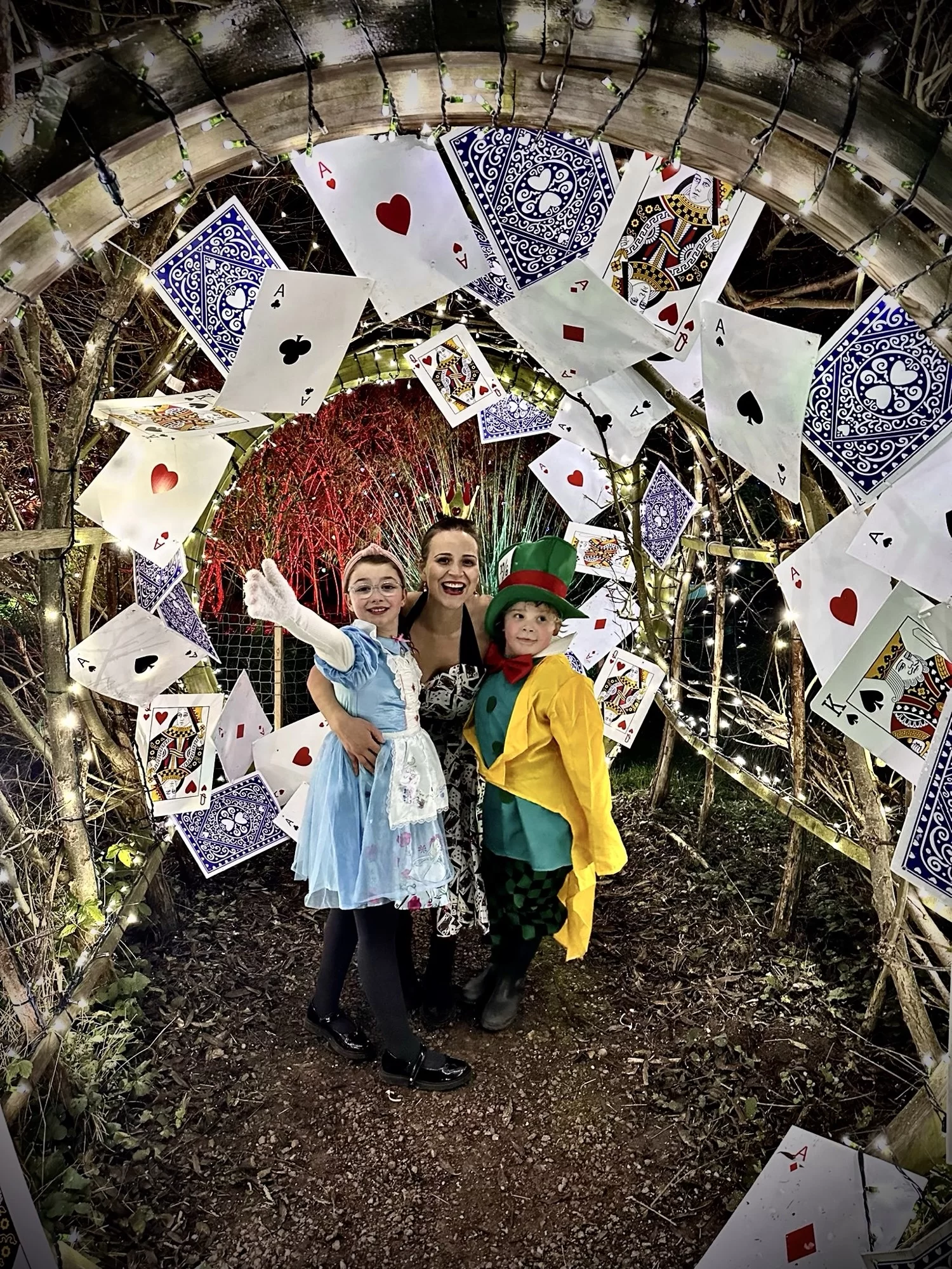 Three people in costumes stand in a tunnel decorated with playing cards and lights.