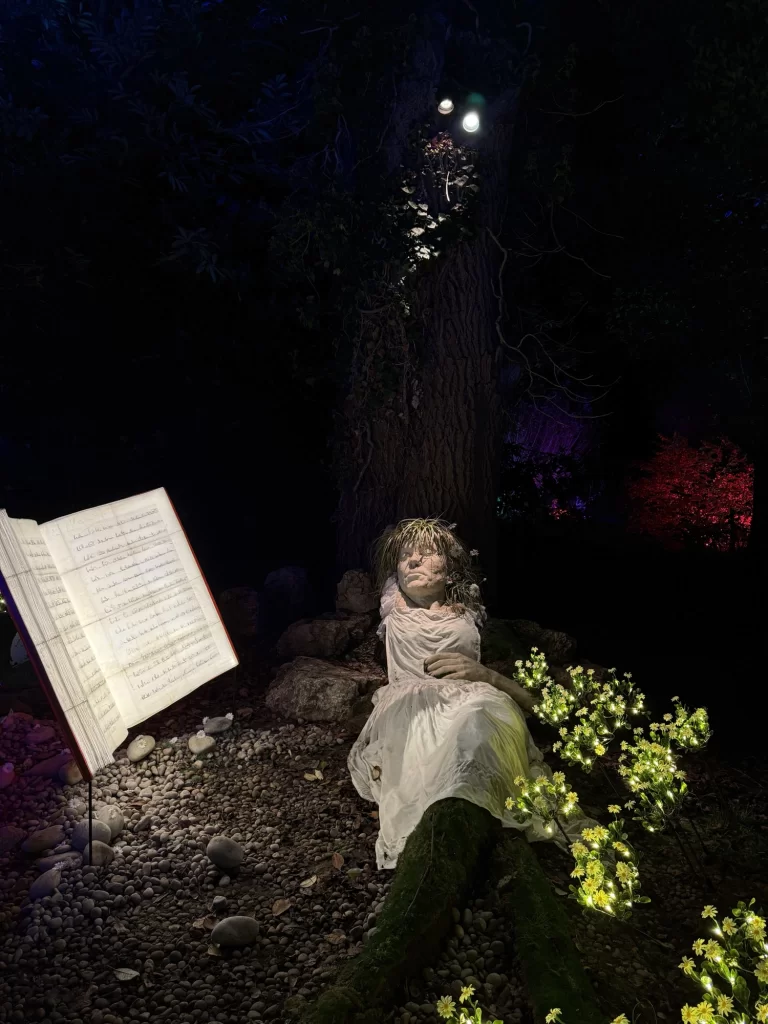 A statue of a person sits against a tree with dim lighting, surrounded by flowers. An open music book is displayed on a stand nearby.