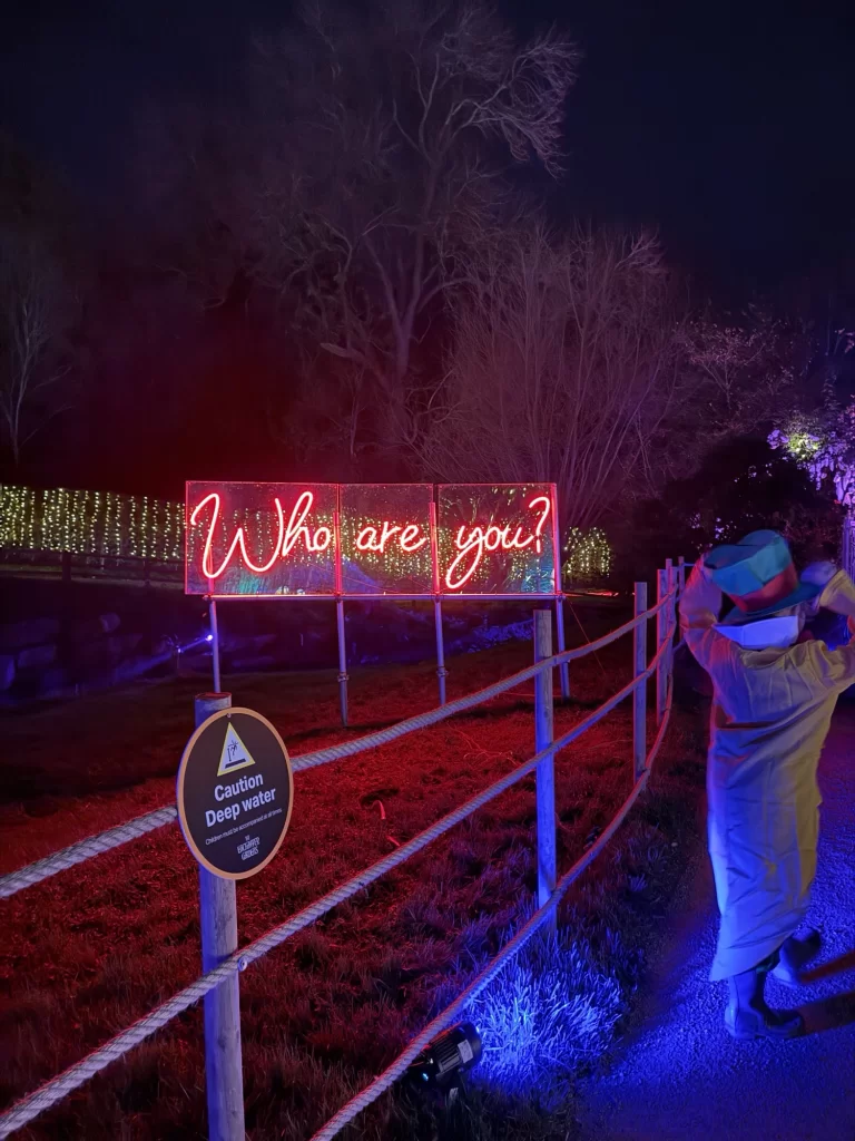 Neon sign reading "Who are you?" illuminated at night. A person in a light-colored outfit stands nearby. A sign warns of deep water.