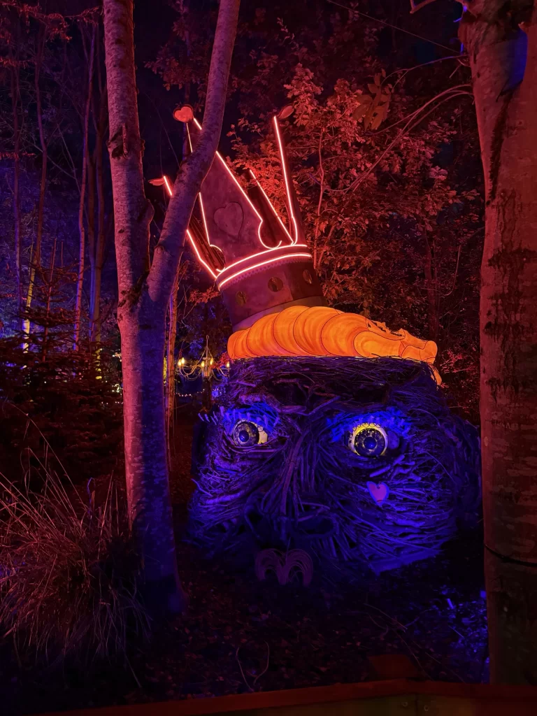 A whimsical outdoor sculpture of a face with glowing eyes and a crown, illuminated by purple and red lights, surrounded by trees.