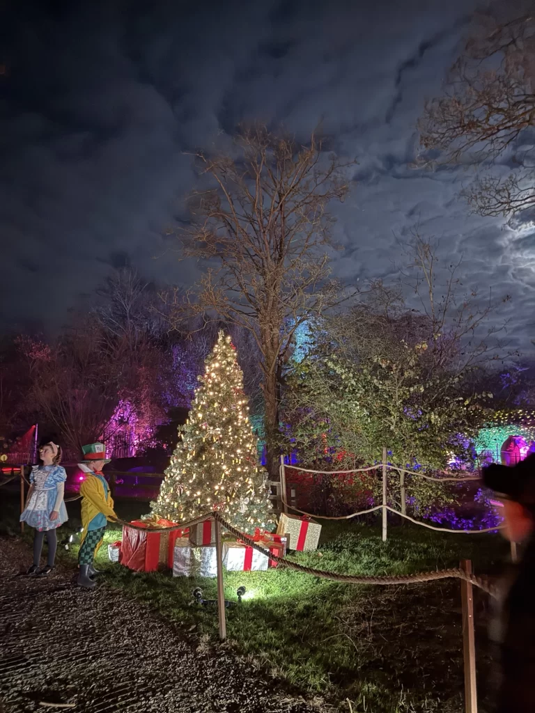 Festive outdoor scene with a lit Christmas tree, wrapped gifts, and two people dressed in costumes under a cloudy night sky.