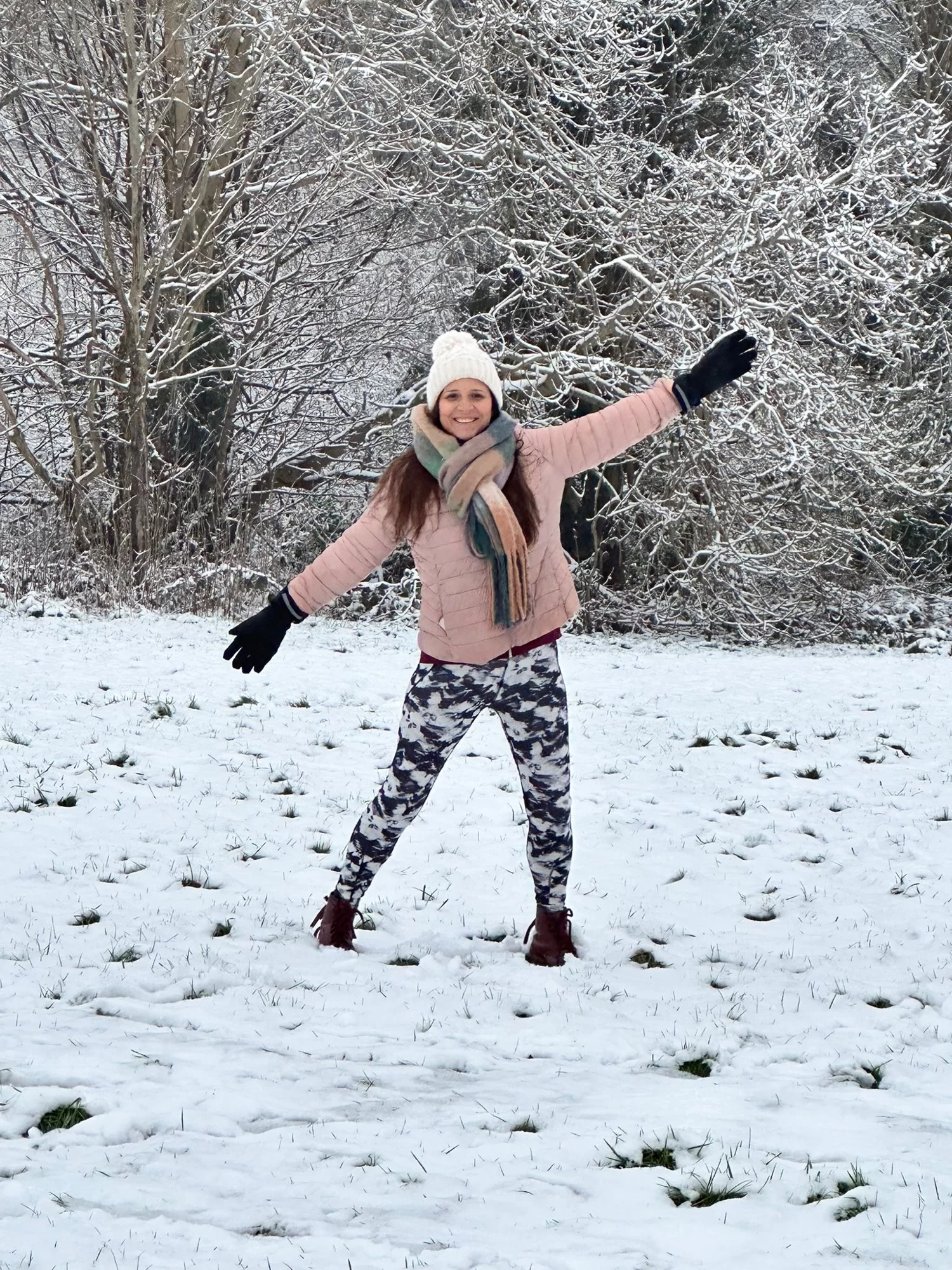 Person smiling and posing in a snowy field, wearing a winter jacket, hat, scarf, and gloves, with trees in the background.