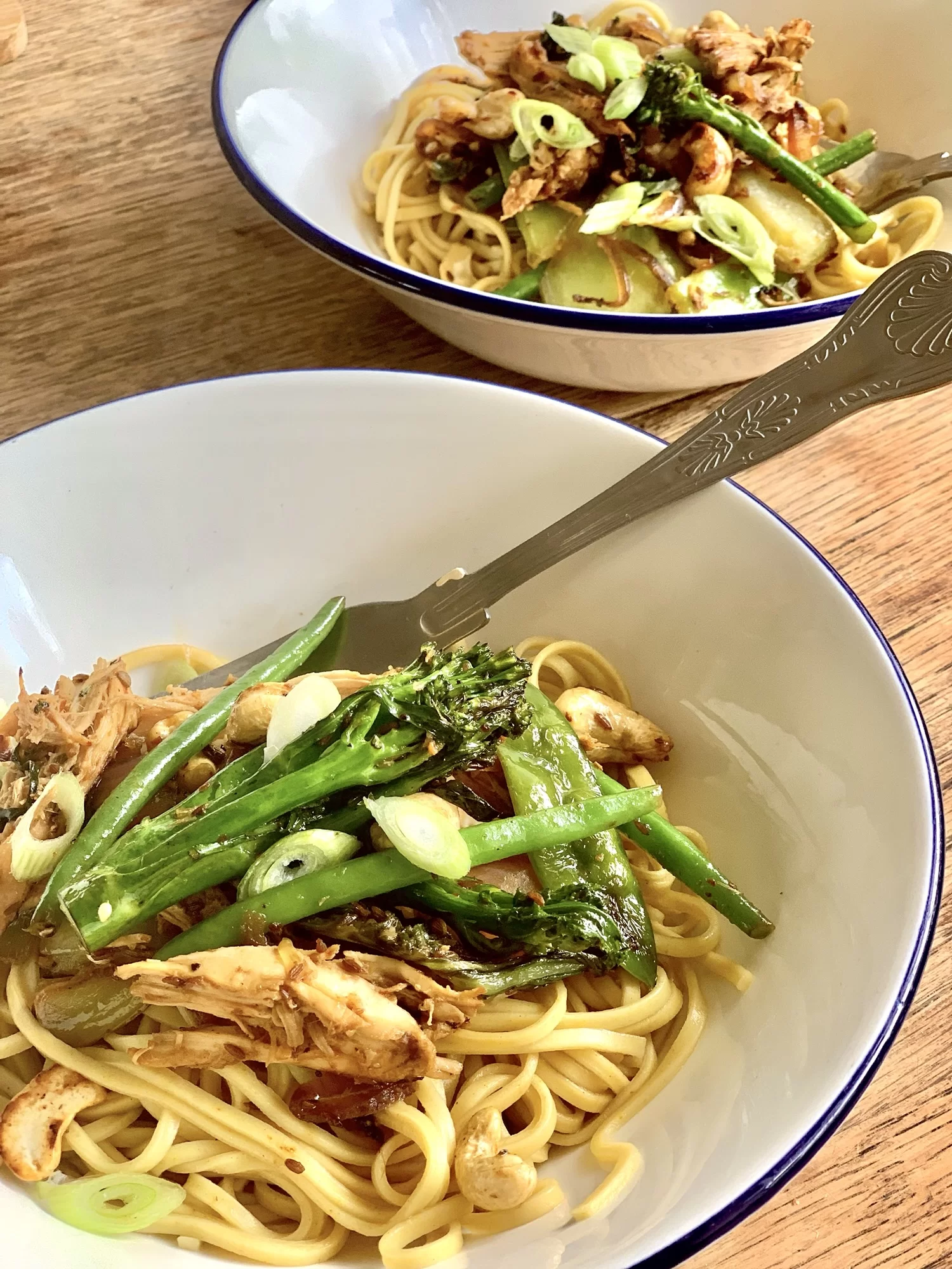 Two bowls of noodles topped with sliced green onions, grilled chicken, and vegetables, with forks resting on the bowl edges.