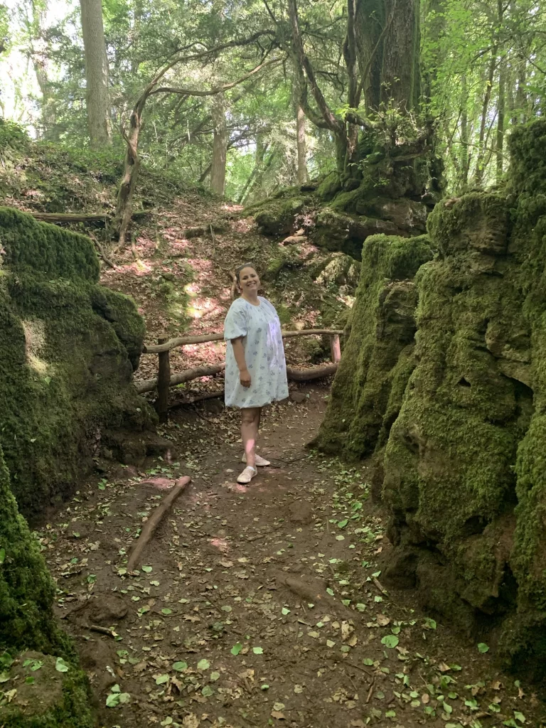 A person in a light dress stands on a wooded path surrounded by moss-covered rocks and trees.