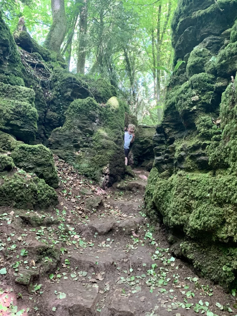 Child peeking from behind moss-covered rock formations on a forest trail.