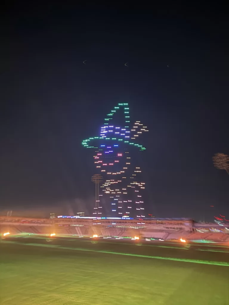 Drones form a colorful image of a smiling witch wearing a hat in the night sky over a stadium.
