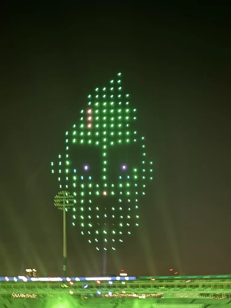 Green and white drones form a face-like pattern in the night sky above a sports stadium.