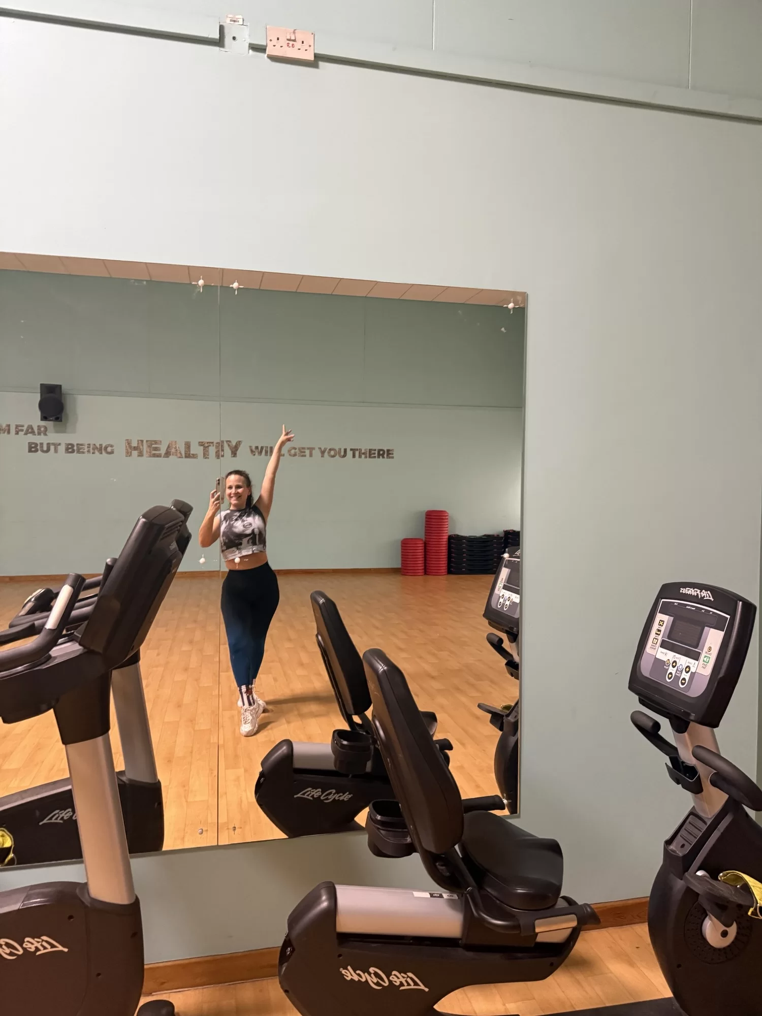 A person posing with an arm raised in a gym mirror, surrounded by exercise bikes. A motivational phrase is partially visible on the wall.