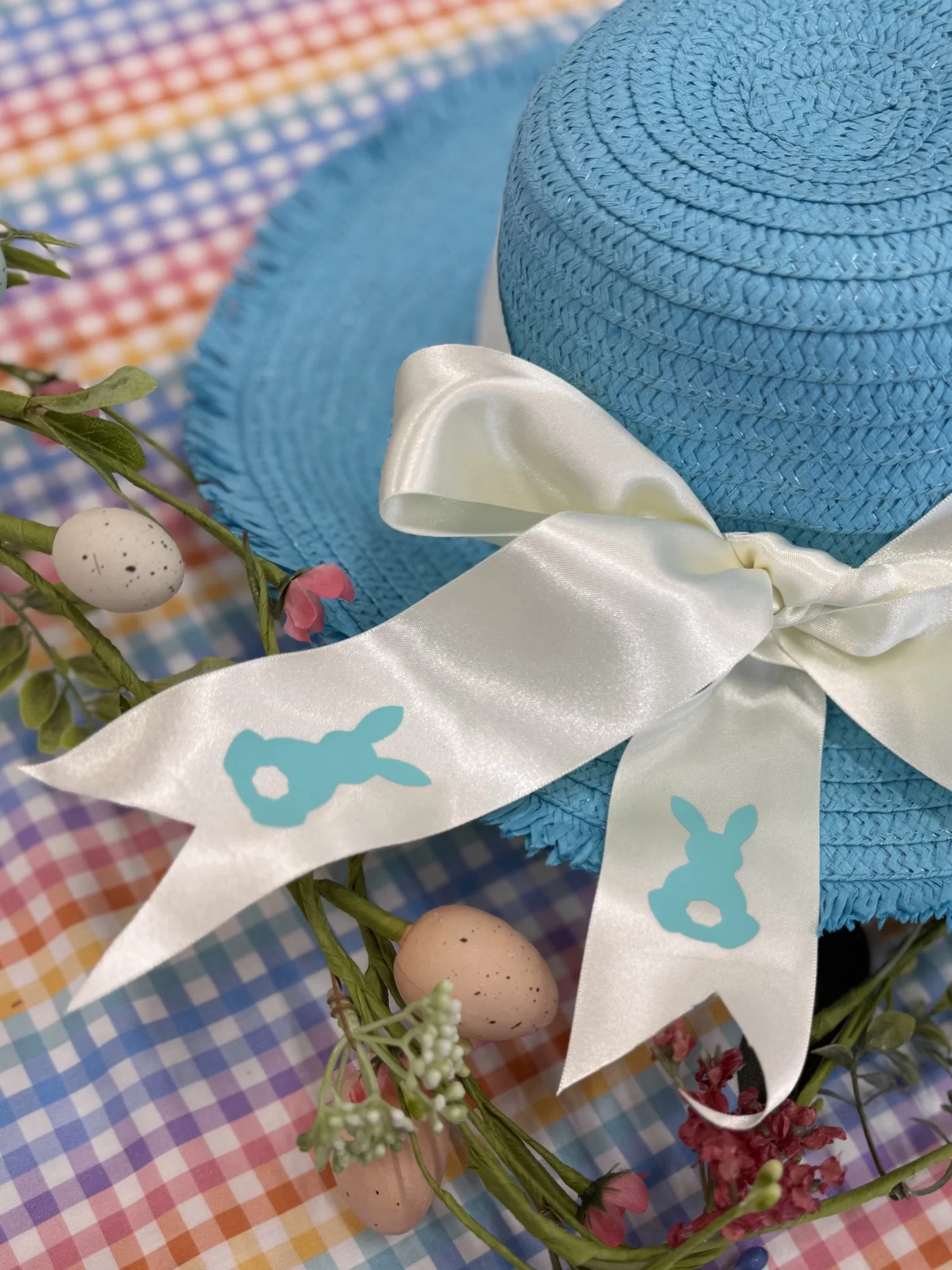 Blue straw hat with white ribbon, featuring blue bunny decorations. Surrounding it are decorative eggs and flowers on a colorful gingham fabric.