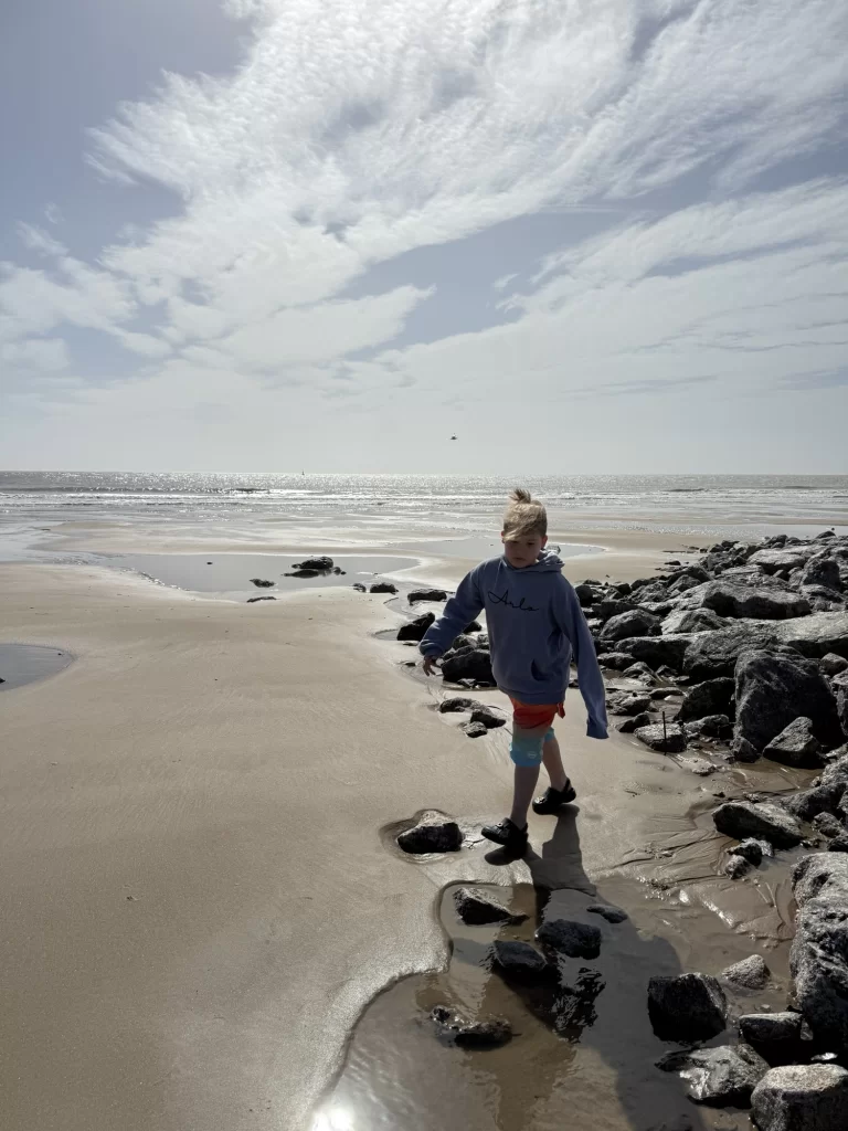 A child in a blue hoodie and red shorts walks on a rocky, sandy beach with the ocean and cloudy sky in the background.