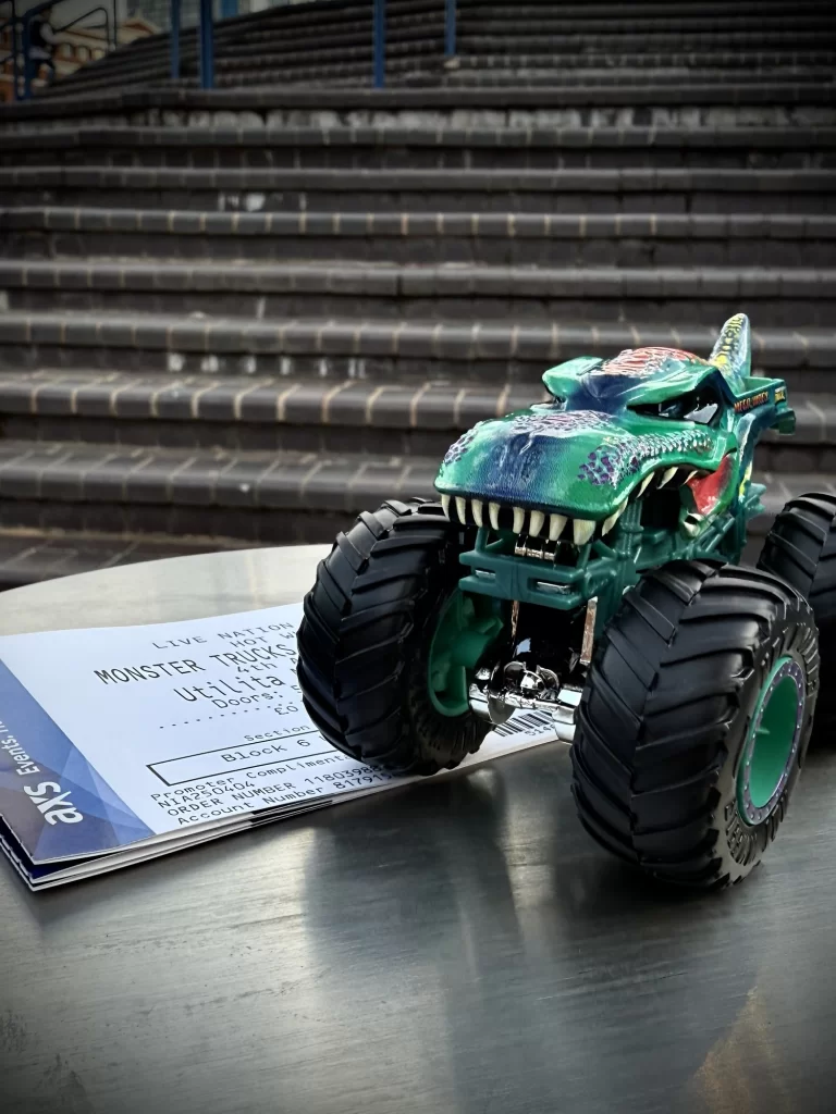 A toy monster truck sits on top of three event tickets on a metal surface. In the background are outdoor steps.