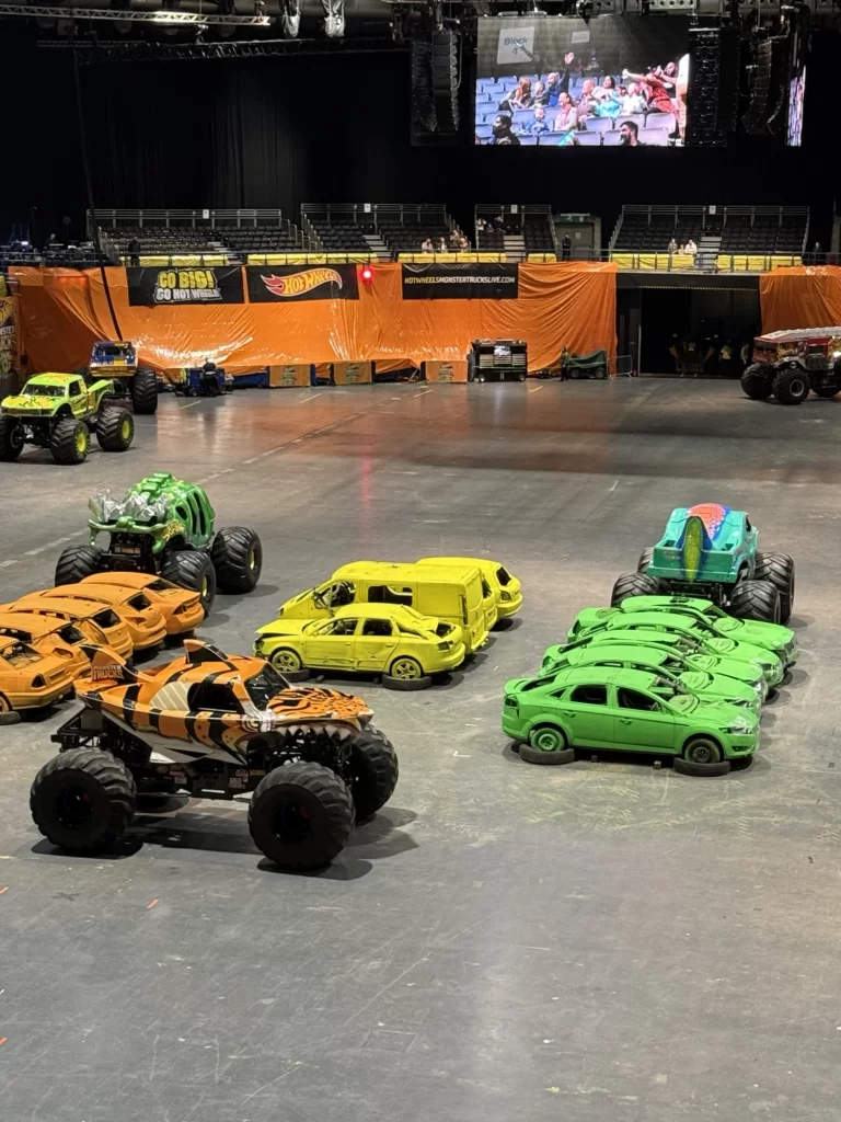 Monster trucks and cars in bright colors are parked inside an arena. Orange banners and a screen displaying a crowd are visible in the background.