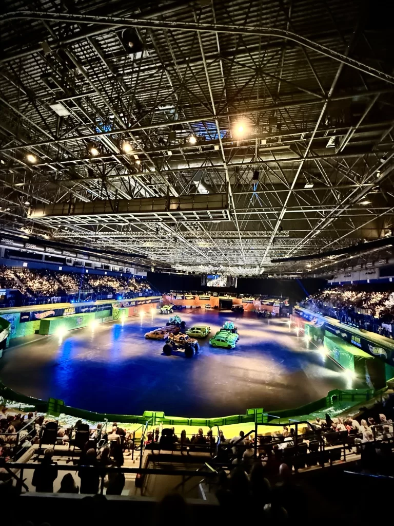 Indoor arena with a large audience, featuring multiple cars on a bright, illuminated track. Lights and metal structures are visible on the ceiling.