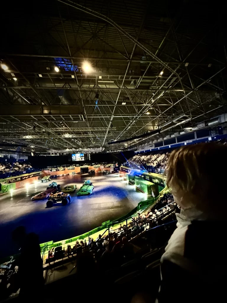Indoor event with vehicles on a lit arena floor, surrounded by tiered seating filled with people, under a large ceiling with industrial lighting.