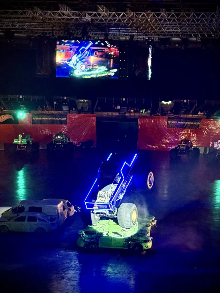 Monster truck performing a backflip in an indoor arena, with neon lights highlighting the truck. A large screen displays the action above the audience seating area.
