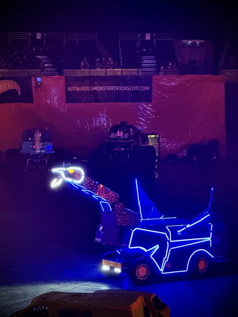 A monster truck show featuring a truck outlined in glowing blue lights, with a dinosaur head attachment, performing in an indoor arena.