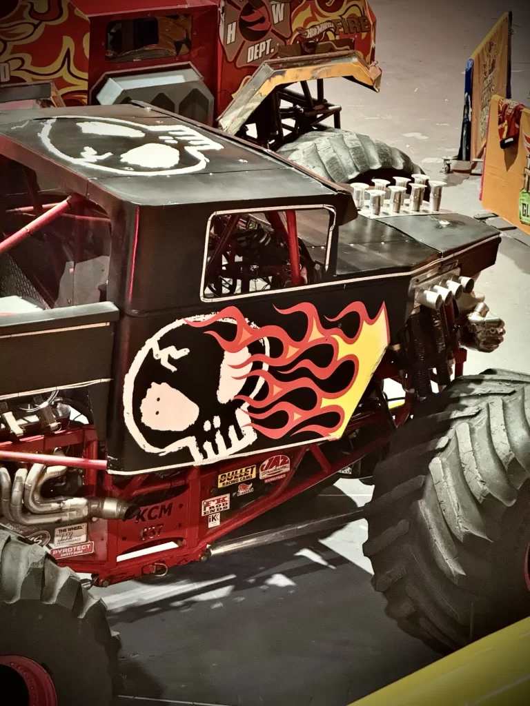 Close-up of a monster truck with large tires, featuring a black body with a skull and flame graphic on the side. Another truck is partially visible in the background.