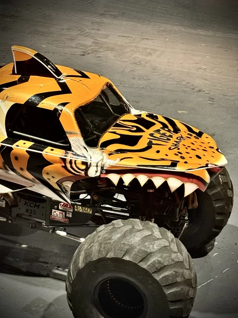 Monster truck with a tiger-themed design, featuring sharp teeth and a striped pattern, parked on a concrete floor.