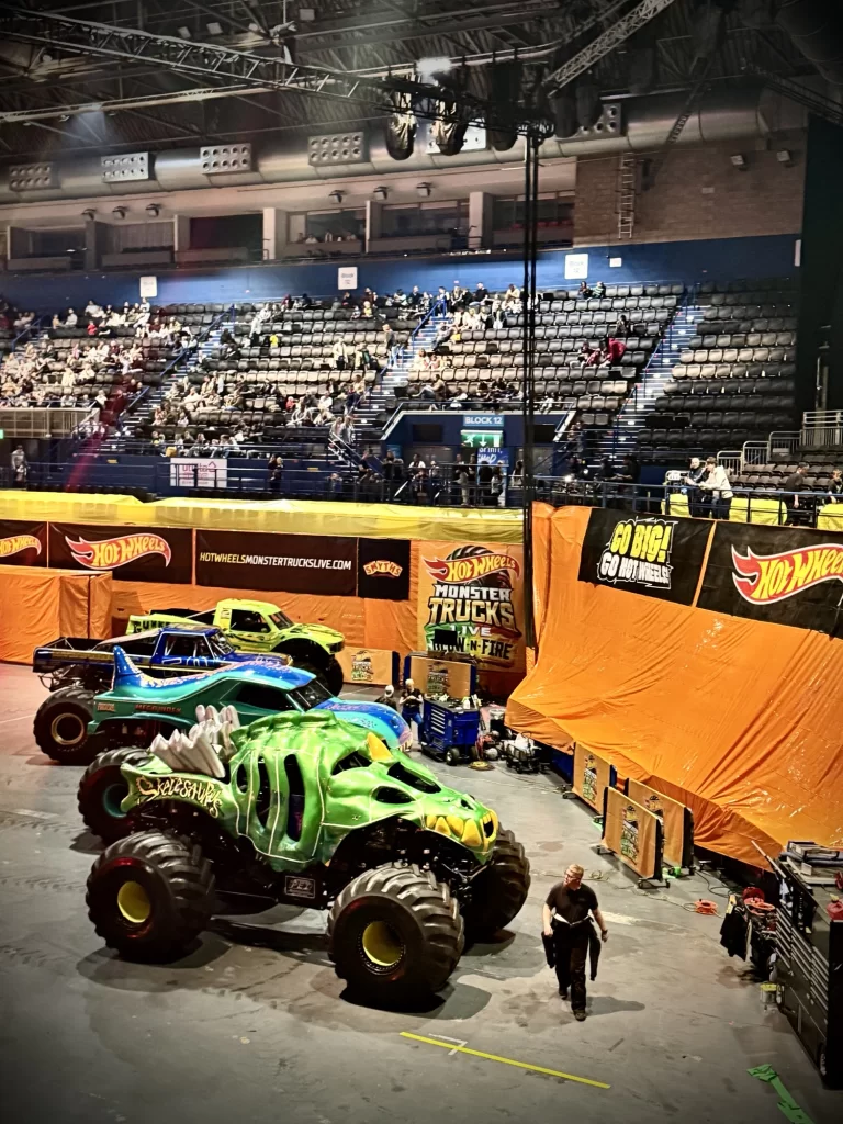 Monster trucks parked indoors with colorful designs, including green and blue. Orange barriers in the background. Sparse audience visible in the stands above.