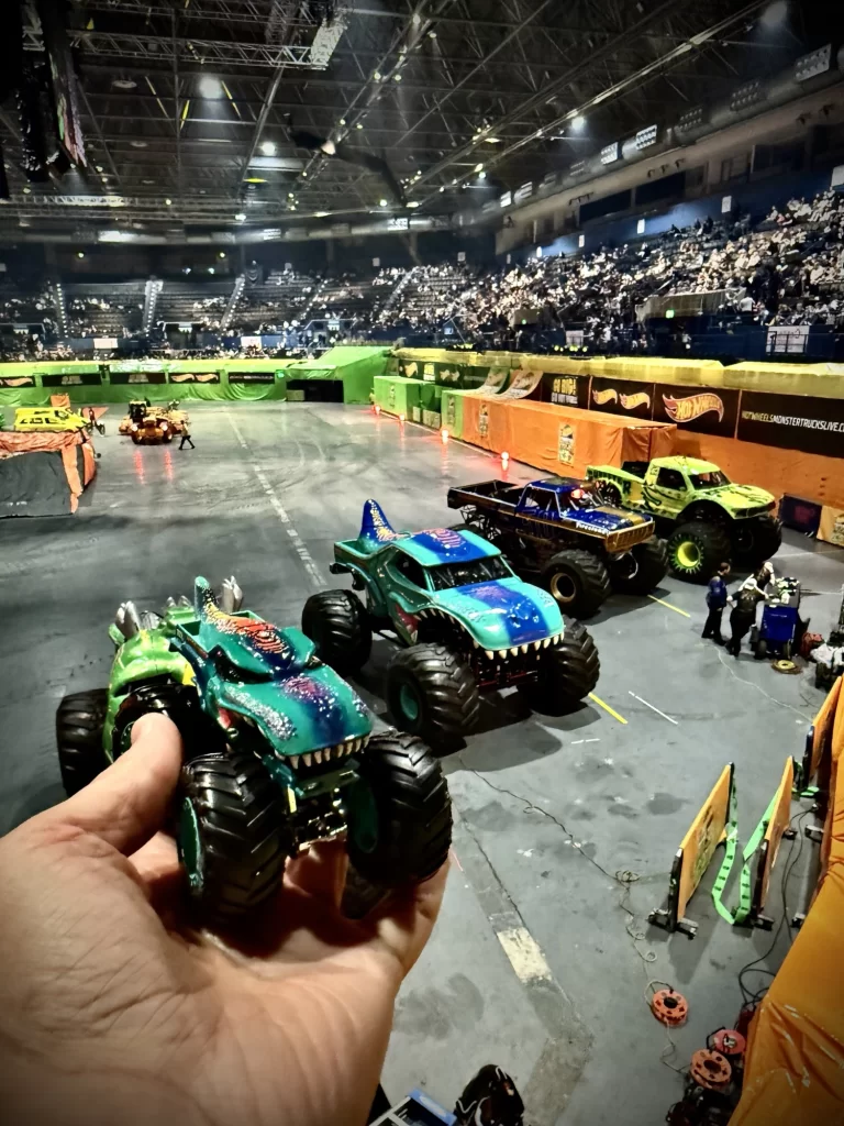 A hand holds toy monster trucks in the foreground. In the background, a monster truck event is taking place in an indoor arena filled with spectators.