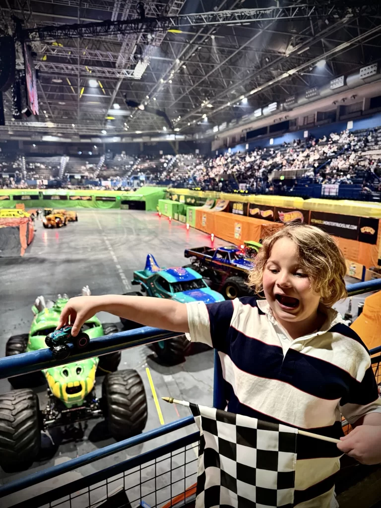 Child holding a toy monster truck and a checkered flag, overlooking a monster truck event in an indoor arena filled with an audience.