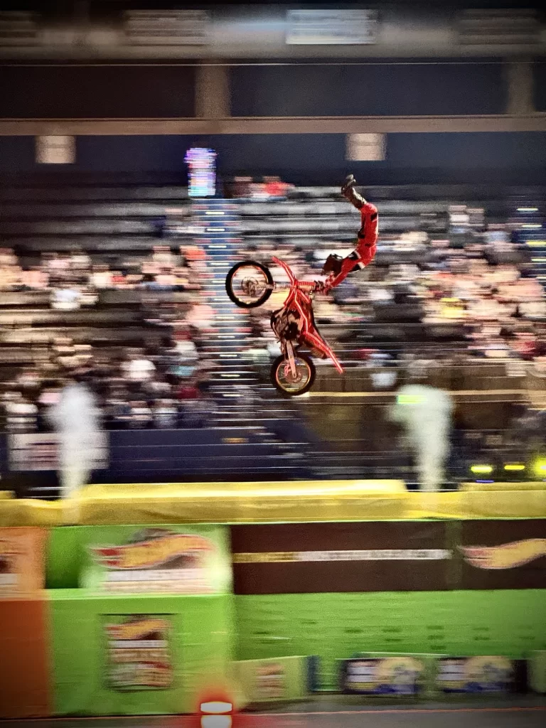 Motorcyclist performing a mid-air stunt in an indoor arena, with blurred crowd in the background, wearing red gear and riding a red bike.