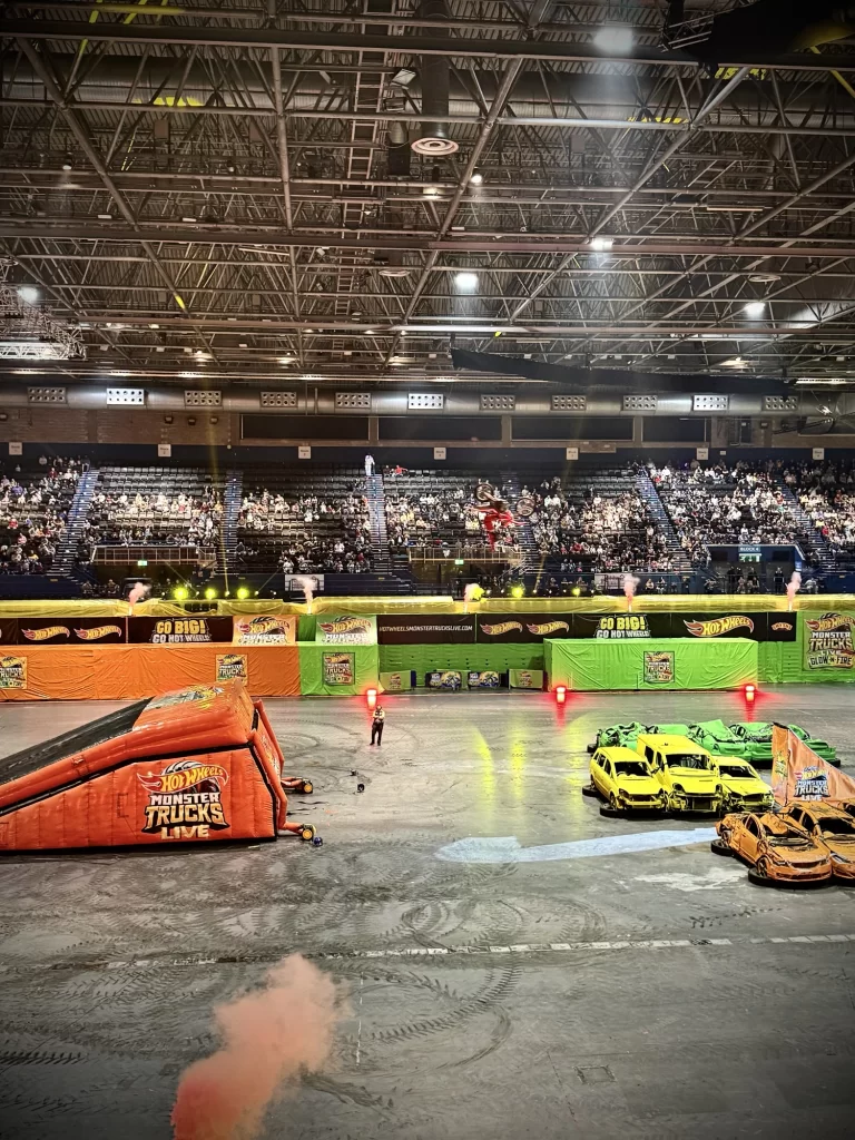 Indoor arena with a Hot Wheels Monster Trucks Live event; ramps, crushed cars, and bright barriers on show, with a performer in the center and a large audience in the stands.