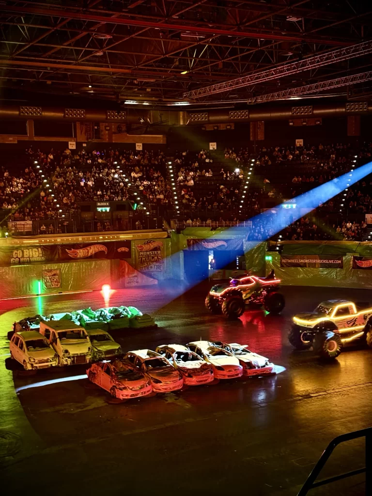 Monster trucks with illuminated frames drive in an arena, surrounded by an audience. Several parked cars are on the floor, and colorful spotlights enhance the scene.
